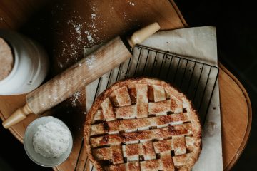 An apple pie on a cooling rack next to a rolling pin and bown of flour