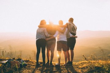 A group of four people standing on a mountain top, with their arms around each others' shoulders and their faces to the sun