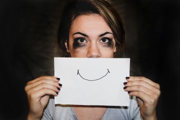 A woman with smudged eye makeup, possibly from crying, holding a picture of a smile in front of her mouth